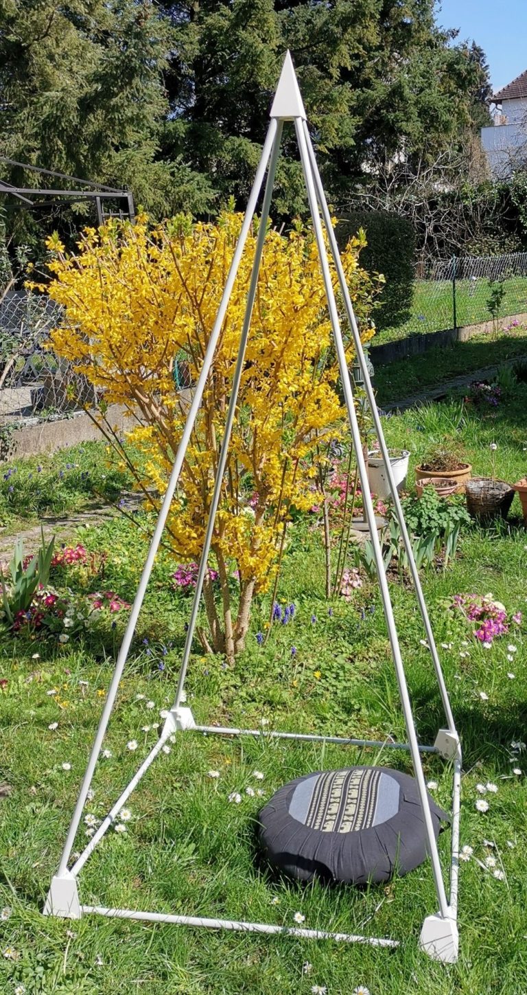 Meditationspyramide Eine Meditationspyramide mit einem Sitzkissen steht in einem Garten mit blühenden Pflanzen.