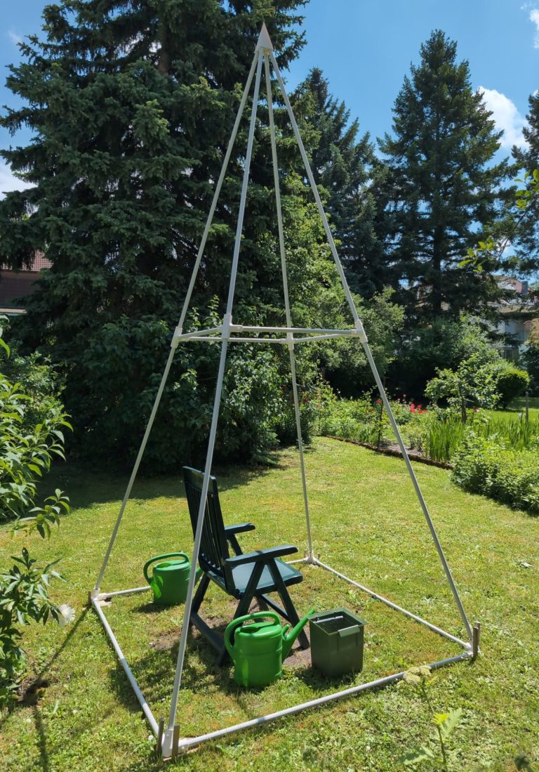 Gartenpyramide Eine große Pyramide die in einem Garten steht. In ihr steht ein Gartenstuhl und Gießkannen.